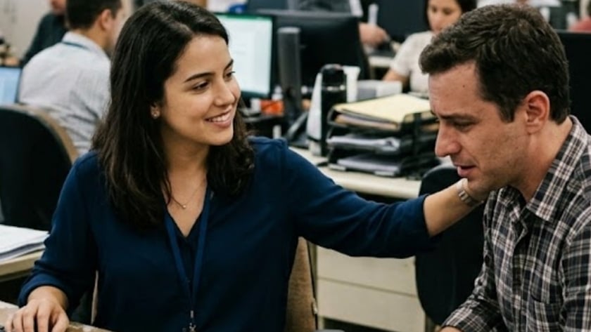 Mulher sorridente com crachá estende o braço em escritório movimentado com colegas ao fundo.