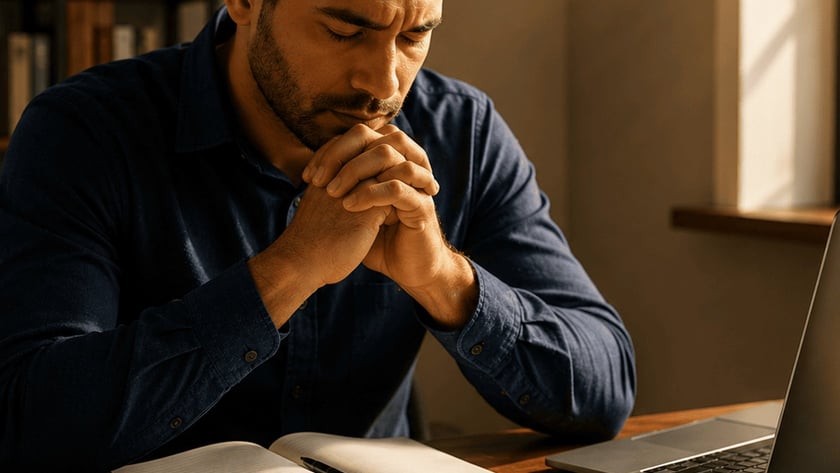 Homem de camisa azul, mãos unidas e olhos fechados, à mesa com notebook, moedas e papéis.