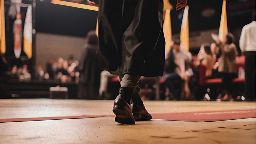 Pessoa de beca preta caminha em um palco de formatura diante da plateia desfocada.