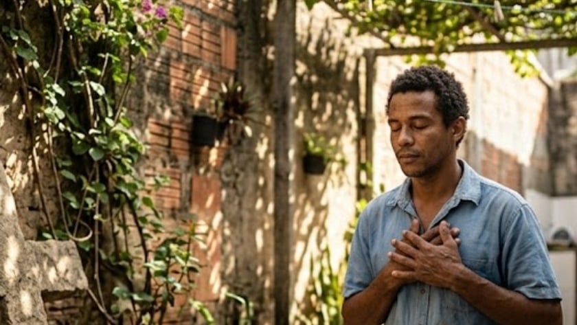 Homem ajoelhado com as mãos no peito e olhos fechados, ao lado de uma cruz de pedra, em um quintal.