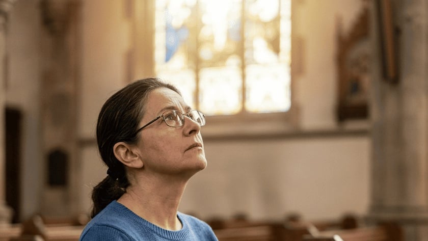 Mulher de óculos e suéter azul, sentada num banco de igreja, olha para cima.