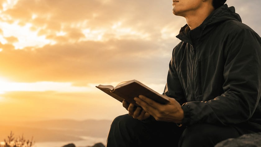 Homem de jaqueta escura sentado em rochas, segurando um livro aberto e olhando para o céu.