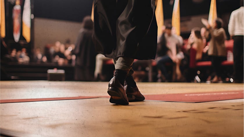 Pessoa de beca preta caminha em um palco de formatura diante da plateia desfocada.