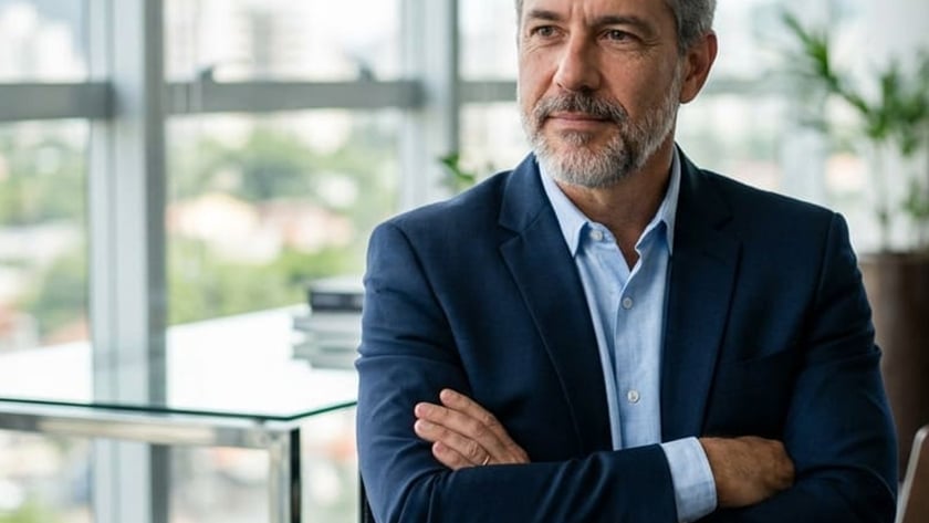 Homem grisalho de blazer azul e braços cruzados ouve outra pessoa em escritório envidraçado.