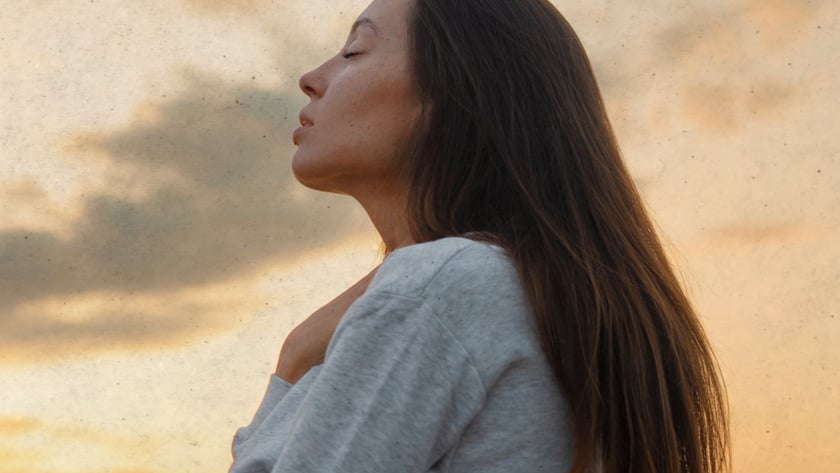 Mulher de perfil, olhos fechados, mão no peito, com moletom cinza ao entardecer.