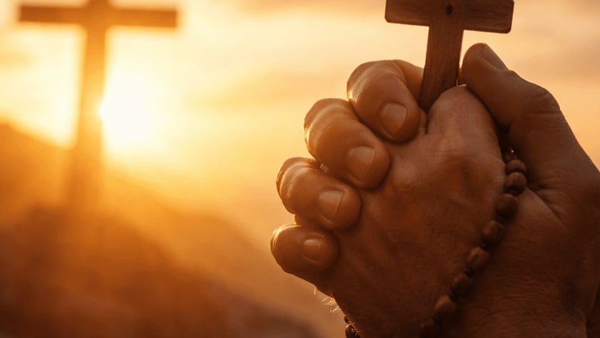 Mãos postas segurando um terço com crucifixo de madeira diante de uma cruz ao pôr do sol.