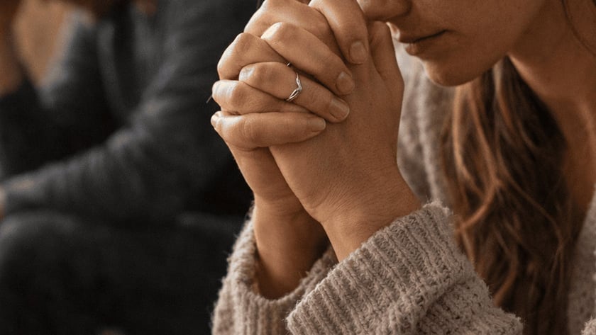 Mulher de mãos unidas e olhos fechados, com homem de cabeça baixa ao fundo.