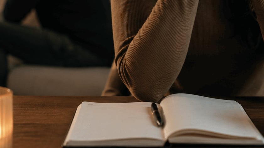 Mulher de mãos unidas diante de um caderno aberto, com homem sentado ao fundo.