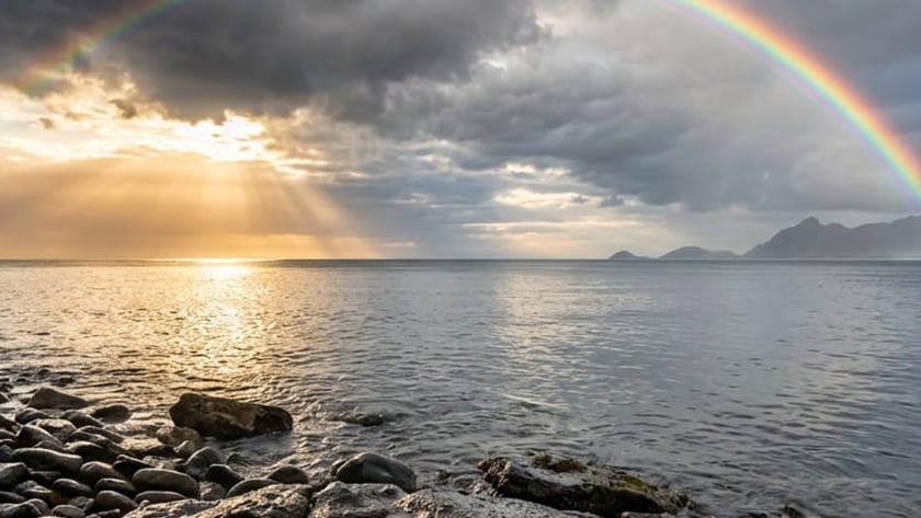 Arco-íris sob nuvens escuras sobre o mar e uma costa de pedras arredondadas.