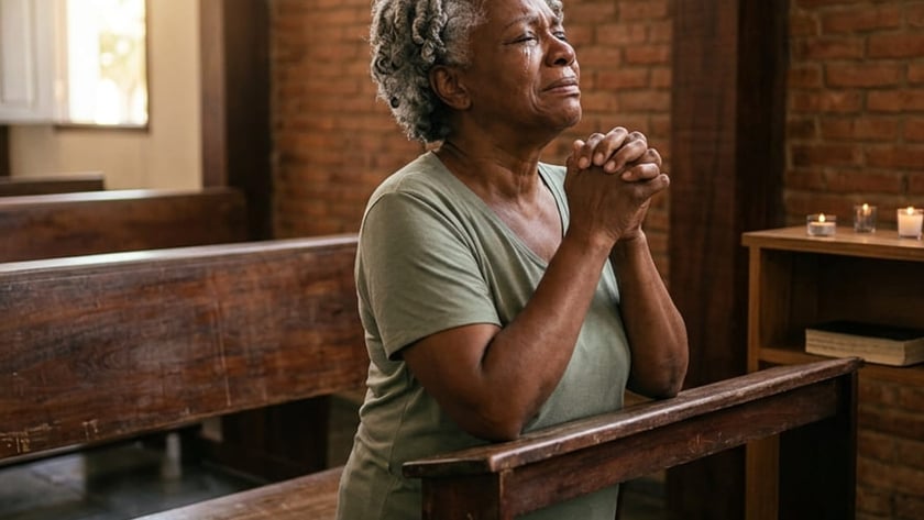 Mulher idosa de cabelos grisalhos ajoelhada, com as mãos unidas, em um banco de igreja.