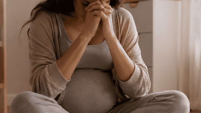 Mulher grávida sentada no tapete, de pernas cruzadas, com as mãos unidas diante do rosto.