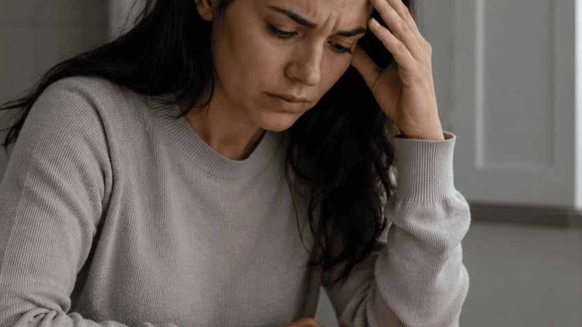 Mulher de cabelo escuro apoia a cabeça na mão à mesa, ao lado de caderno e roupas dobradas.