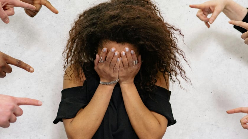 Mulher de cabelo cacheado cobre o rosto com as mãos, cercada por dedos apontados.