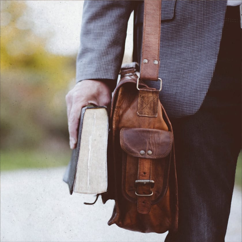 Homem de paletó segura um livro grosso e carrega uma bolsa de couro marrom.