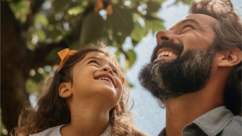 Homem barbudo e menina de cabelo cacheado olham para cima, sorrindo, sob uma árvore.