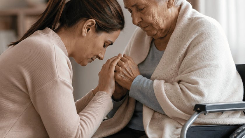 Mulher ajoelhada segura as mãos de uma idosa em cadeira de rodas, em uma sala.