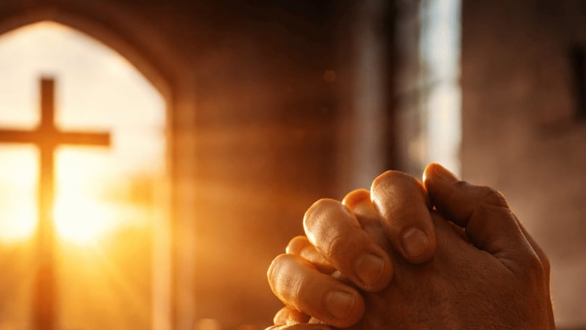 Mãos postas ao lado de uma Bíblia aberta, diante de uma cruz contra a janela.