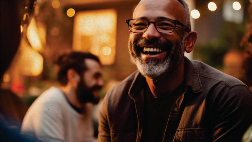 Homem de óculos e barba grisalha rindo em um jardim com luzes ao fundo.