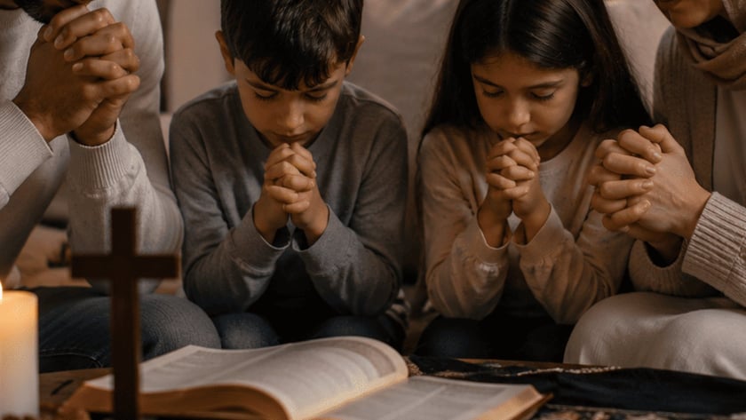 Homem, menino e menina rezam de mãos juntas diante de uma cruz, velas e Bíblia.