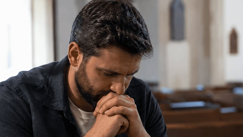 Homem de barba, com as mãos unidas junto ao rosto, sentado em um banco de igreja.