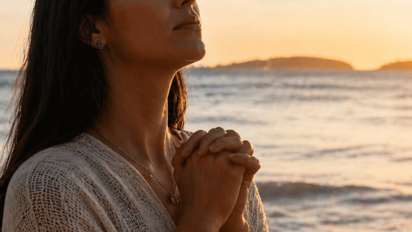 Mulher de cabelos escuros e olhos fechados, com as mãos unidas, na praia.