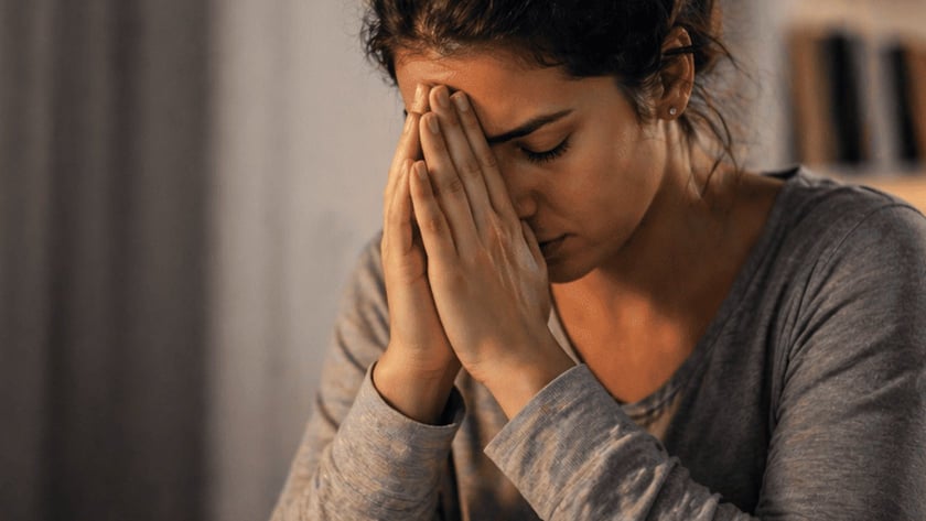 Mulher de coque e blusa cinza, sentada à mesa, esconde o rosto entre as mãos juntas.