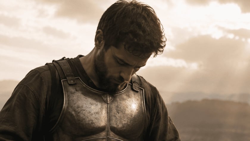 Homem barbado ajoelhado afivela o cinto de uma armadura de metal em um morro.