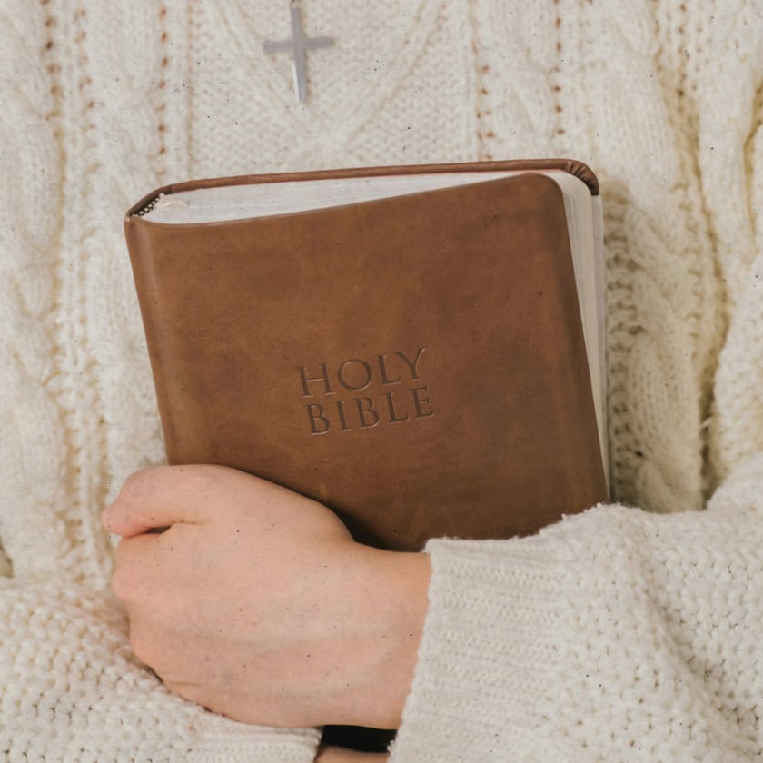 Pessoa de suéter de tricô branco segura uma Bíblia marrom junto ao peito: "HOLY BIBLE".