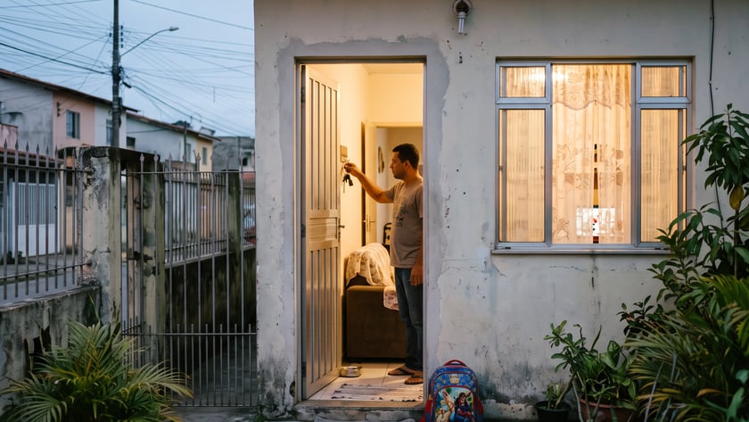 Homem na porta de casa simples ao entardecer, com chaves na mão e mochila no chão.