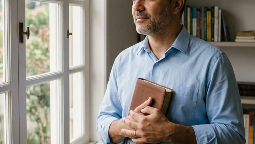 Homem grisalho de camisa azul segura um livro marrom e olha pela janela.
