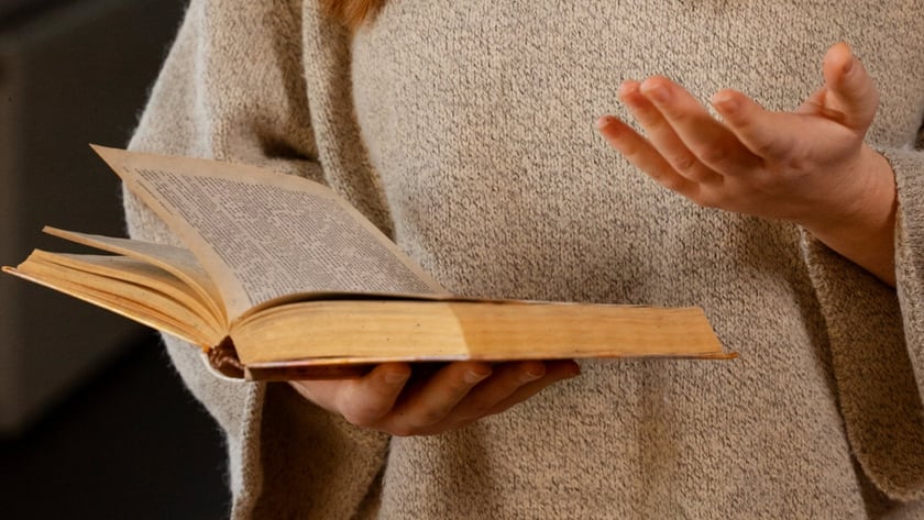 Mulher de cabelo ruivo e suéter bege segura um livro antigo aberto e gesticula.
