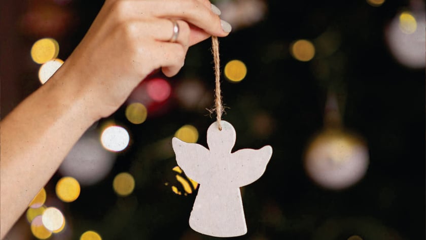 Mão segura enfeite de anjo branco pendurado por barbante diante de árvore de Natal.