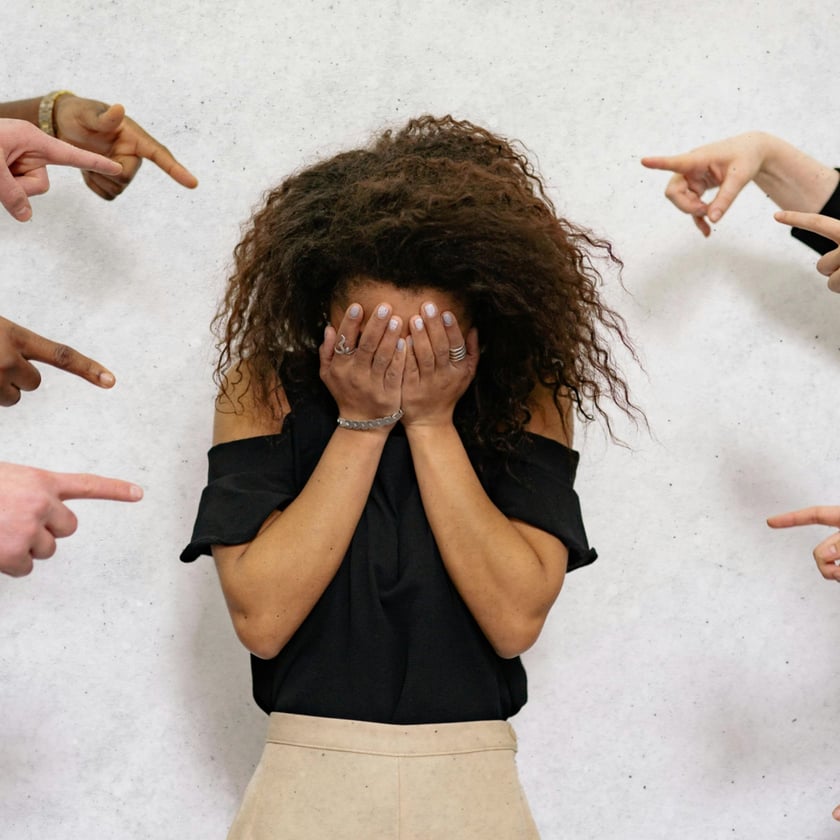 Mulher de cabelo cacheado cobre o rosto com as mãos, cercada por dedos apontados.