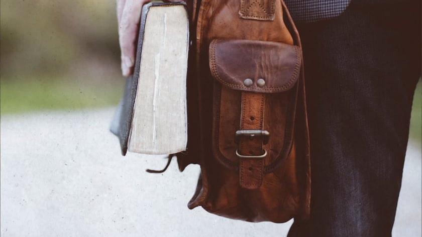 Homem de paletó segura um livro grosso e carrega uma bolsa de couro marrom.