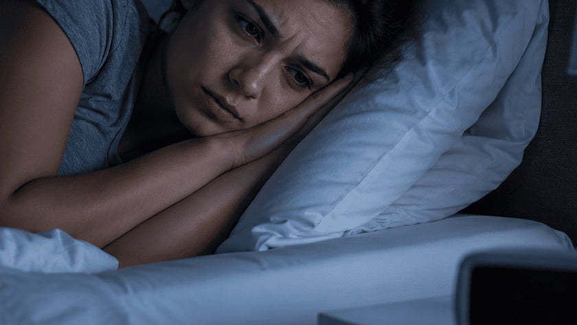 Mulher deitada na cama, de olhos abertos, olhando para o despertador na mesa de cabeceira.