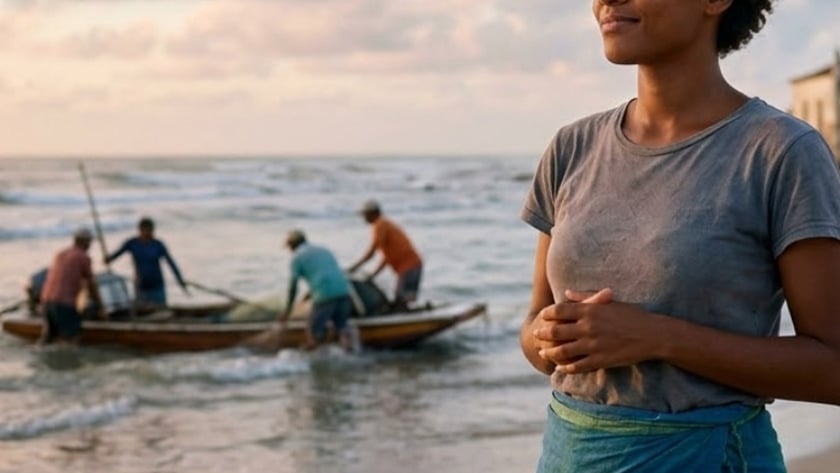Mulher de cabelo curto em pé na praia, com pescadores em um barco ao fundo.