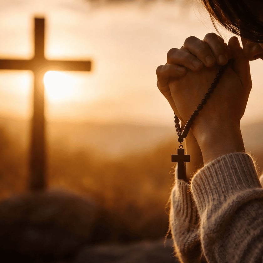 Mulher de suéter claro, de cabeça baixa, reza com um terço nas mãos diante de uma cruz.