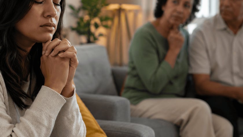 Mulher de olhos fechados e mãos juntas no sofá, com um casal de idosos ao fundo.