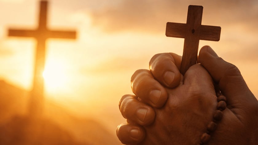 Mãos postas segurando um terço com crucifixo de madeira diante de uma cruz ao pôr do sol.