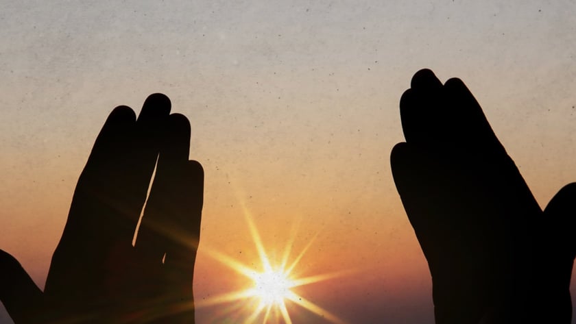 Duas mãos erguidas em silhueta enquadram o sol brilhando no horizonte ao entardecer.