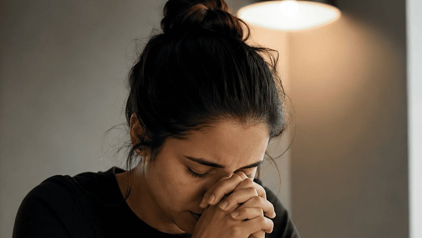 Mulher de coque e blusa preta, de olhos fechados e mãos unidas junto ao rosto