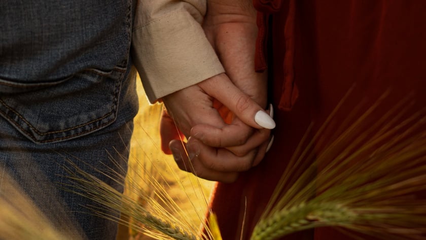 Duas pessoas de mãos dadas em um campo de trigo, uma de jeans e outra de vestido vermelho.