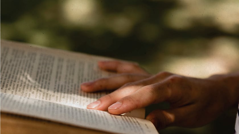 Mãos de uma pessoa segurando um livro aberto ao ar livre, com os dedos sobre a página.