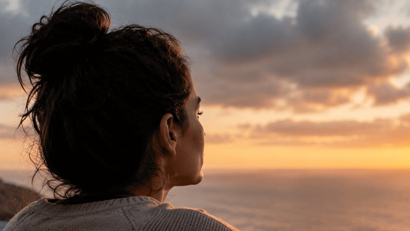Mulher de coque e suéter bege, vista de costas, observa o mar do alto de um penhasco.