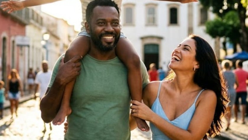 Homem carrega menina de braços abertos nos ombros, ao lado de mulher, em rua de paralelepípedos.
