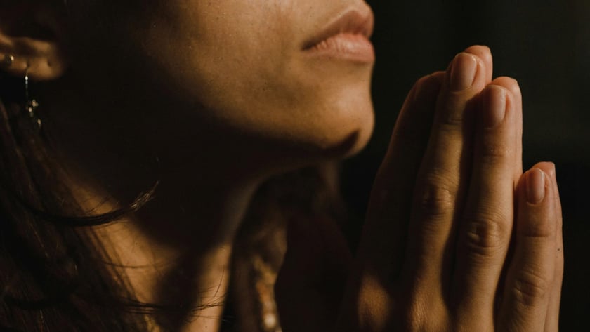 Mulher de cabelos escuros com as mãos unidas diante do queixo, em um ambiente escuro.