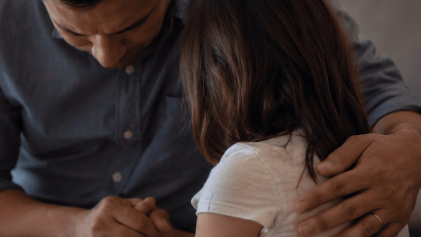 Homem de camisa azul-escura abraça uma menina de cabelo castanho, de mãos dadas, dentro de casa.