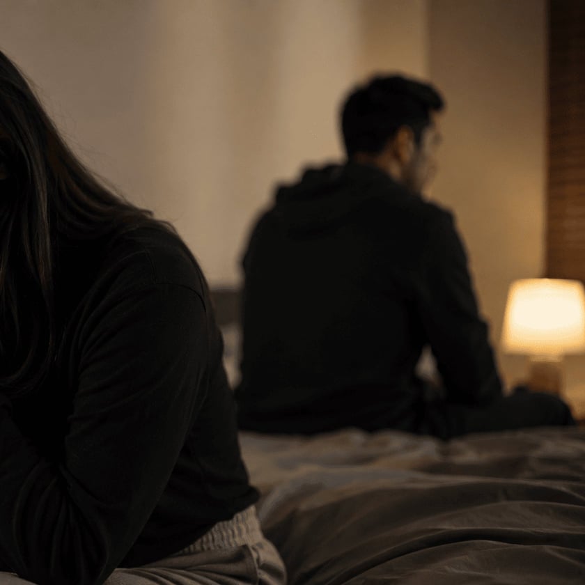 Mulher de cabelo longo e homem sentam em lados opostos da cama, de costas um para o outro.