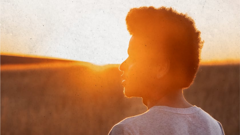 Jovem de camiseta clara, visto de costas e de perfil, olha um campo ao pôr do sol.