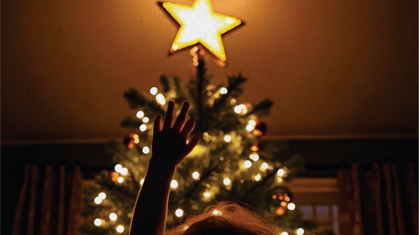 Criança de costas estica a mão em direção à estrela de uma árvore de Natal.